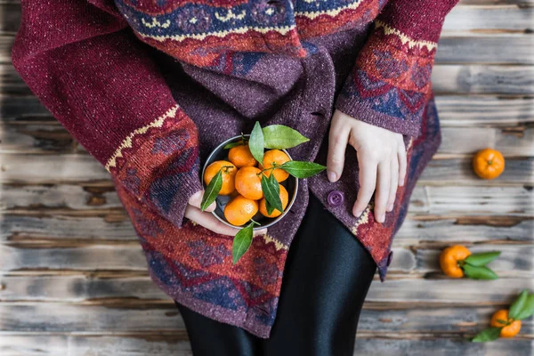 Woman in a huge winter sweater sits on the wooden rustic floor with small iron bucket of tangerines