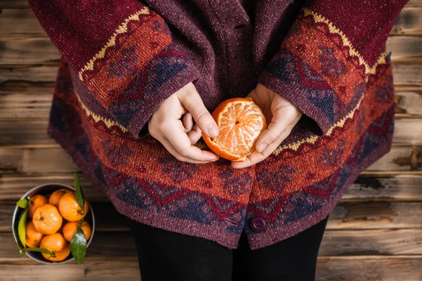 Woman in a huge winter sweater sits on the wooden rustic floor and cleaning the mandarin