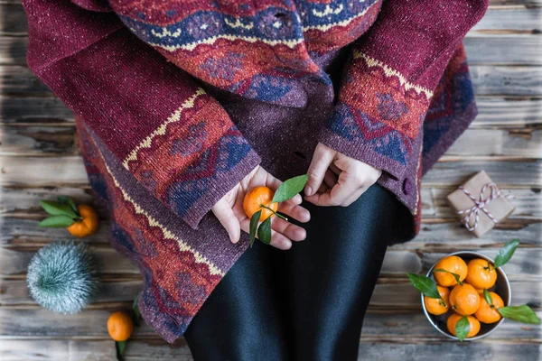 Woman in a huge winter sweater sits on the wooden rustic floor with christmas gifts and mandarins