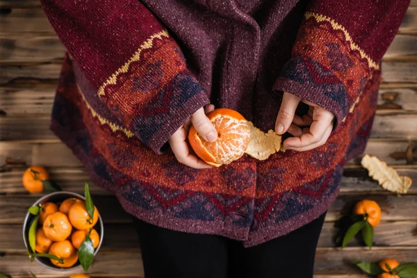 Woman in a huge winter sweater sits on the wooden rustic floor and cleaning the mandarin