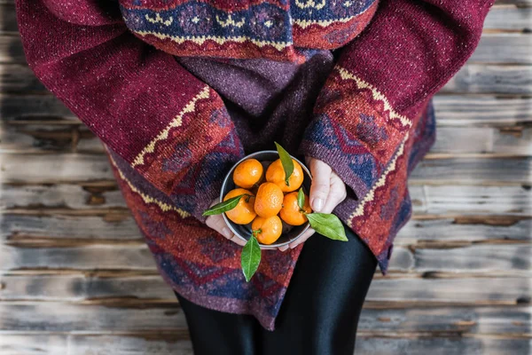 Woman in a huge winter sweater sits on the wooden rustic floor with small iron bucket of tangerines