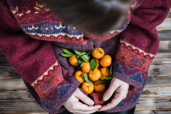 Woman in a huge winter sweater sits on the wooden rustic floor with handful of mandarins in her hands