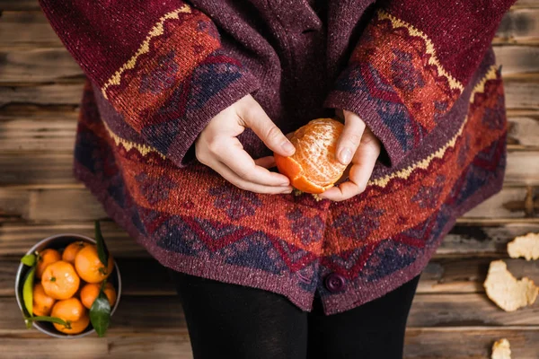 Woman in a huge winter sweater sits on the wooden rustic floor and cleaning the mandarin