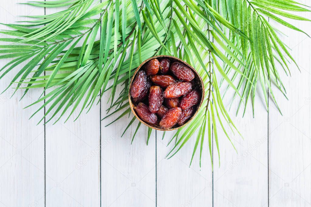 Royal dates fruit in a bowl of coconut in palm branches and leaves on a white wooden table. 