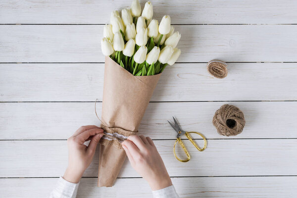 Woman florist wrapping beautiful bouquet of white tulips in pack craft paper on the wooden table. Flat lay