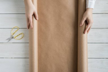 Dişi eller, tahta beyaz masaya hediye ve çiçek paketlemek için bir rulo kahverengi el işi kağıt açıyor. Boşluğu kopyala. 