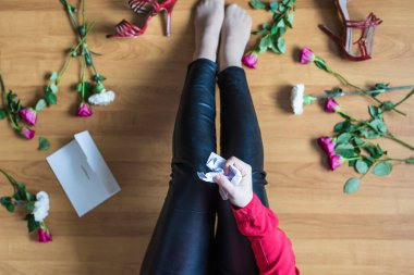 Unhappy woman sitting on the floor among scattered flowers and reading letter. Broken relationships in bad Valentine's day concept. Top view