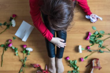 Unhappy woman sitting on the floor among scattered flowers and reading letter. Broken relationships in bad Valentine's day concept. Top view
