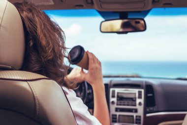 Kadın sürücü araba kullanırken arabada kağıt bardak içiyor. Arka planda okyanus tropik plajı boyunca araba gezisinde dinlenen bir kız. Gezgin konsepti. Arka plan