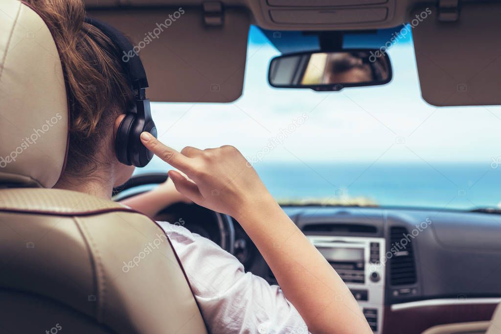 Woman driver in the headphones driving a car. Girl relaxing in auto trip traveling along ocean tropical beach in background. Traveler concept. Back view