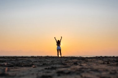 Denizin karşısındaki sahilde duran mutlu kadın silueti ve yükselen güneş. Yaz ve özgürlük kavramı. Güneş ışınları (ışınlar) dişilerin bacaklarında parlıyor.