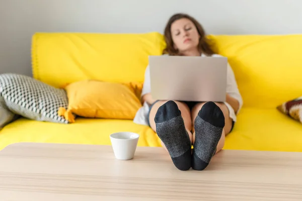 Woman freelancer sitting on the couch with laptops in the socks and putting legs on the table. Work at home. Concept. Cup coffee. Relax at home.