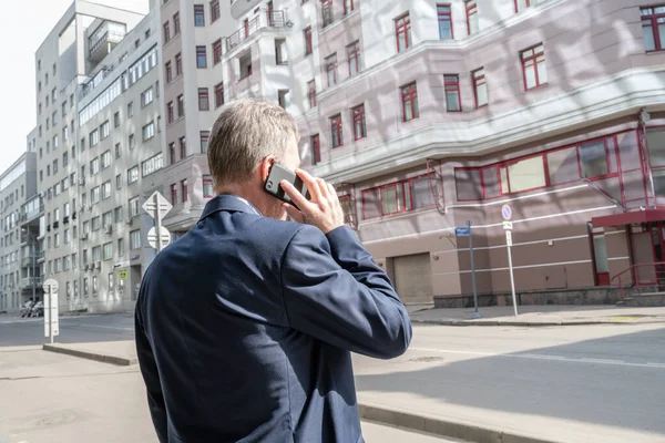 Alone middle-aged gray-haired businessman in a blue suit calling by phone standing on the empty city street.
