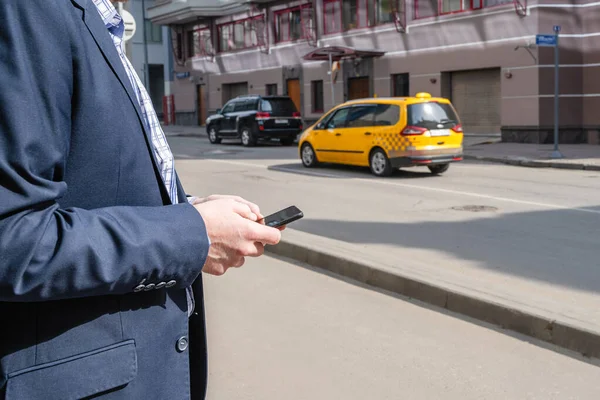 Businessman in a suit booking a taxi using mobile phone app standing on the street. yellow taxi car on the background of office buildings of the finance business center. Concept. Taxi city service