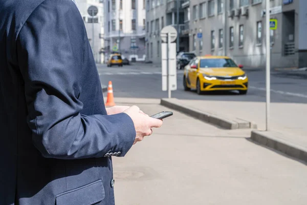 Businessman in a suit booking a taxi using mobile phone app standing on the street. yellow taxi car on the background of office buildings of the finance business center. Concept. Taxi city service