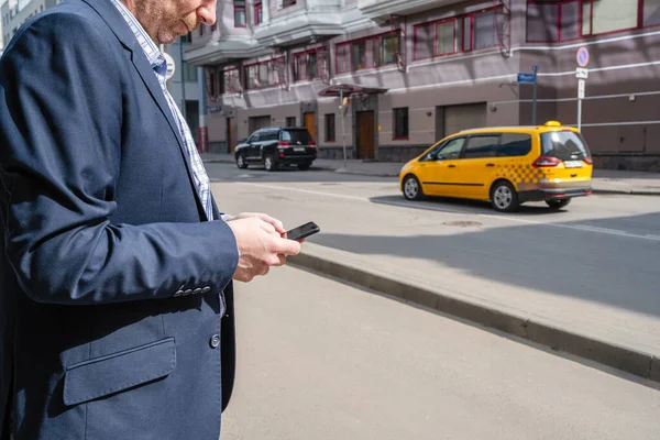 Businessman in a suit booking a taxi using mobile phone app standing on the street. yellow taxi car on the background of office buildings of the finance business center. Concept. Taxi city service