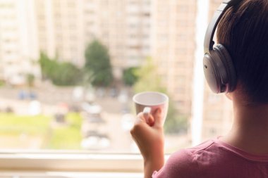 Evinin penceresinde duran ve kulaklıkla müzik dinleyen yalnız bir genç kadının arka planına. Kız sabahın köründe kahve içer..