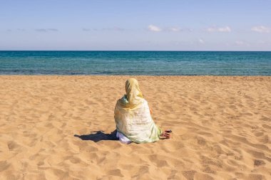 Renkli bir başörtüsü takmış, kumsalda kumsalda oturan ve denize bakan yalnız Müslüman kadın yolcuların arka planı..