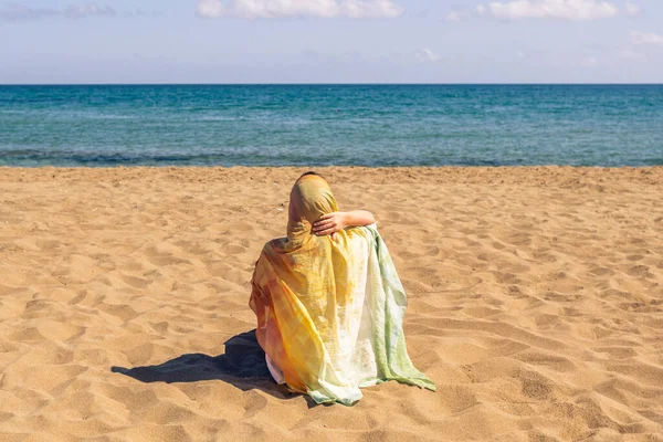 Renkli bir başörtüsü takmış, kumsalda kumsalda oturan ve denize bakan yalnız Müslüman kadın yolcuların arka planı..