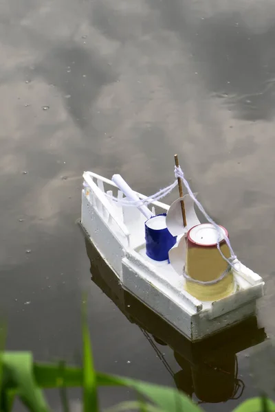 The white boat floats on the water, next to the green reeds. A makeshift boat is drifting on a pond near the shore. The sky is reflected in the water. Image of a toy boat on the river.