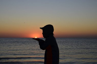 Deniz kıyısında güneşi elinde tutan bir çocuk var. Gün ağarırken sahilde elinde güneş olan genç bir çocuğun silueti. Çocuk yana doğru duruyor ve güneşe bakıyor. Rüya gibi, romantik bir görüntü..