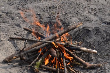 Ateş kumlu sahilde yanıyor. Ağaçların dalları ve dalları ateşte, kumda yanıyor. Ahşap parlak bir ateş alevi ile bir ateşte yatar. Kavram: açık hava eğlencesi, rahatlama.