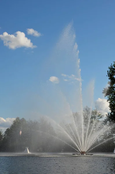 Fountain on the river against the background of large, green trees. Big jets of water, splashing against a blue sky with clouds. Summer and spring landscape. Beautiful landscape of a warm spring day.