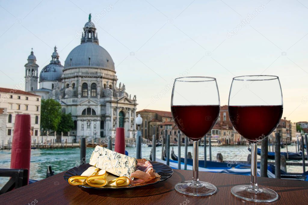 Two glasses of red wine with charcuterie assortment on gondolas and Santa Maria della Salute. Glass of red wine with different snacks - plate with ham, sliced, blue cheese. Couple romantic celebration