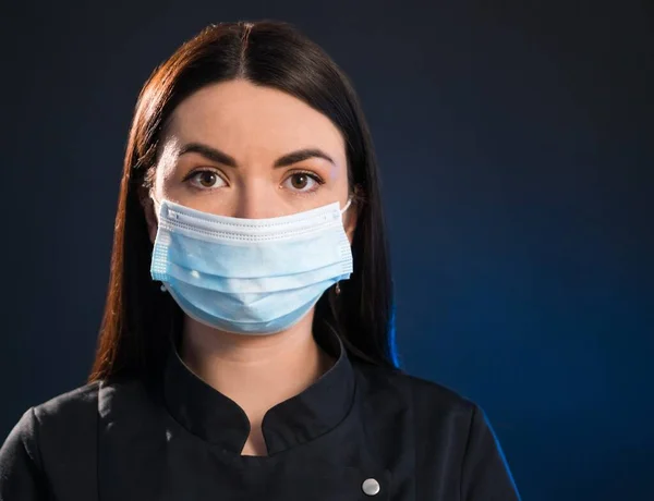 Coronavirus, maskeli kadın. Siyah arka planda, yüz maskesi takan, kameraya bakan genç bir kadının stüdyo portresi. Ayakların jesti. Grip salgını, toz alerjisi, virüs koruması. Şehir hava polisi