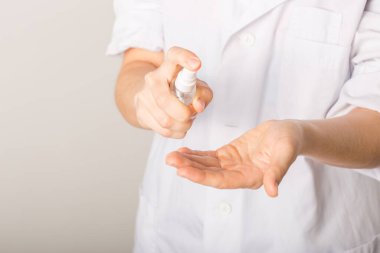 Beyaz önlüklü, beyaz elbiseli, beyaz bir kadın ellerini şişedeki el dezenfektanı ile dezenfekte ediyor.