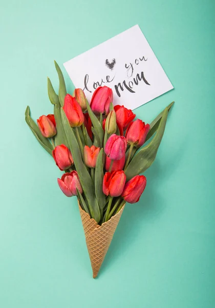 red  beautiful tulips in an ice cream waffle cone with card For  you on a color blue background. Conceptual idea of a flower gift. Spring mood