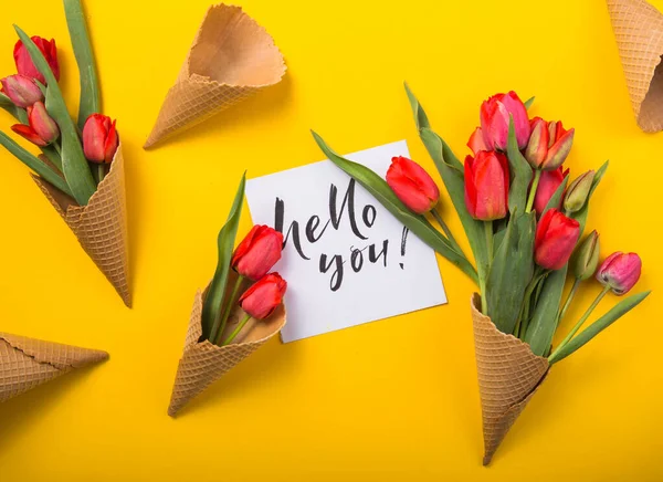 red  beautiful tulips in an ice cream waffle cone  with ink card a yellow color background. Conceptual idea of a flower gift. Spring mood