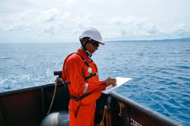 Güvenlik subayı, bir sanayi gemisinde duruyor, tulum, kask, güvenlik gözlüğü takıyor ve kontrol listesi olan bir pano tutuyor..