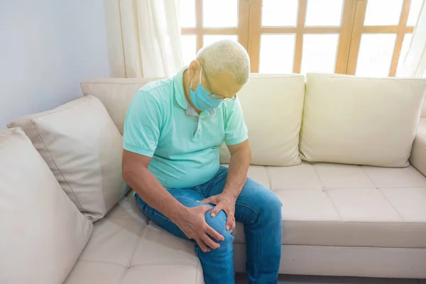 Evde diz ağrısı çeken tıbbi maskeli yaşlı bir adam.