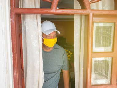 Coronavirus. Pencereden dışarı bakan ve evdeki hastalıktan korunma ve iyileşme maskesi takan hasta bir adam..