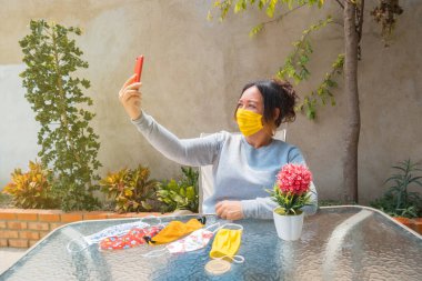 Yüz maskeli bir kadın evde oturmuş cep telefonuyla fotoğraf çekiyor.