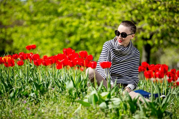 Jonge vrouw zittend op het gras onder de rode tulpen weide in het voorjaar genieten van zonnige dag — Stockfoto