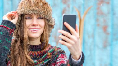  Güzel bir kış kadını, ahşap duvarın üzerinde kazak ve kürk şapka takarak akıllı telefon kullanıyor.