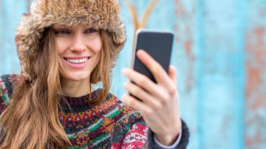  Güzel bir kış kadını, ahşap duvarın üzerinde kazak ve kürk şapka takarak akıllı telefon kullanıyor.