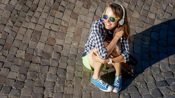 Young woman sitting on paving stones enjoying the sunshine and listening music, tourist girl in bright glasses relaxing outdoors, full length, copy space, view from above