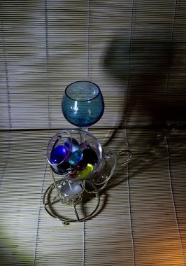 Farklı renklerde cam ve kristal tabaklar, yeşil, mavi, şeffaf şarap bardakları ve masa üzerinde peçeteler ve çerçevelerle renkli cam toplarla şamdanlar.