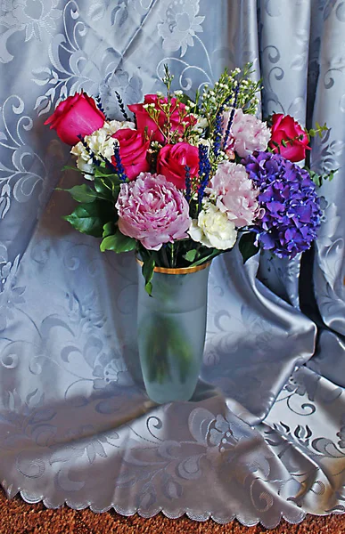 Masanın üzerindeki vazoda bir buket, örtülü mavi arka plan, beyaz şakayık, leylak rengi ve mavi lavanta rengi güller.