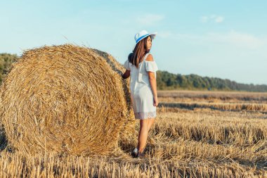 Tarlada saman balyasının yanında duran beyaz elbiseli ve şapkalı çekici genç bir kadın.