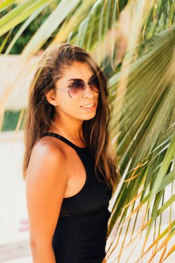 Yaz mevsiminde palmiye yapraklarının yanında duran mayo ve güneş gözlüklü çekici, genç bir kadın.