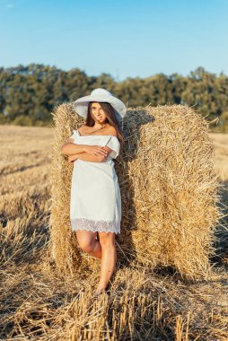 Beyaz elbiseli, şapkalı, saman balyasının yanında poz veren güzel, gülümseyen bir kadın.