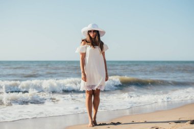Beyaz elbiseli, şapkalı ve güneş gözlüklü, kumlu sahilde yürüyen çekici, genç bir kadın.