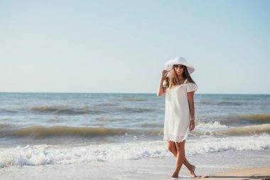 Beyaz elbiseli, şapkalı ve güneş gözlüklü, kumlu sahilde yürüyen çekici, genç bir kadın.