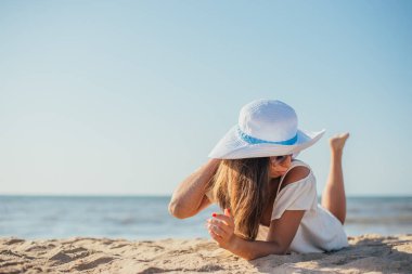 Yazın kumlu sahilde uzanan şapkalı ve güneş gözlüklü çekici bir genç kadın. 