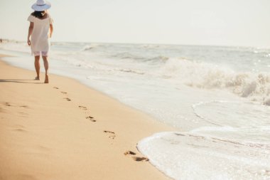 Beyaz elbiseli ve şapkalı genç bir kadının kumsalda deniz kenarında yürüyüşünün arka görüntüsü.