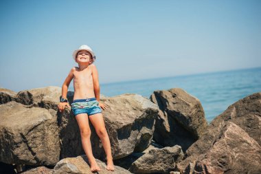 Yaz zamanı kumlu sahilde şortlu ve şapkalı sevimli çocuk oturuyor. 